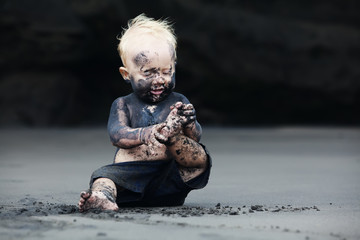 Funny portrait of smiling child with dirty face sitting and playing with fun on black sand sea...