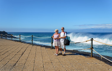 Mature couple is standing on embankment on splashing wave backgr
