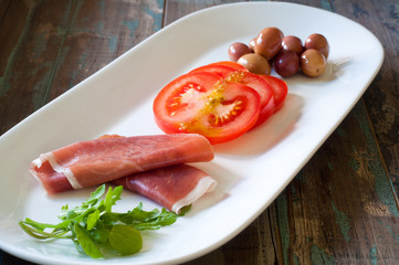 Parma ham, sliced tomato, fresh olives and salad served on a rustic wooden table. A traditional mediterranean snack or side dish.