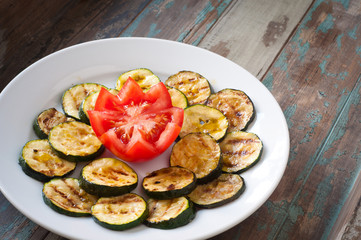 Simple vegetable dish of sliced, grilled courgette / zucchini served with fresh tomato on a rustic wooden table.
