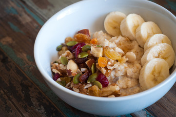 Bowl of freshly prepared home made porridge oatmeal topped with sliced banana and dried fruit, nuts and seeds. A healthy breakfast served on a rustic wooden tray.