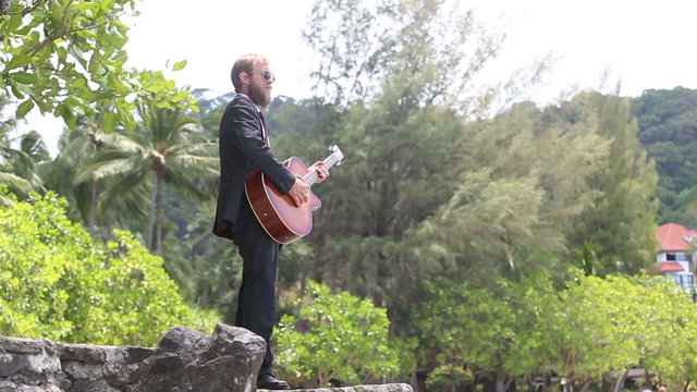 guitarist in black gesticulates actively against tropical trees	