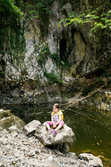 Pensive little girl sitting on a big rock by a small pond