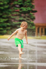 Portrait of happy toddler boy jumping in puddles during the rain thunderstorm on a bright summer day outside, sports recreation leisure concept, childhood and freedom, freeze motion effect
