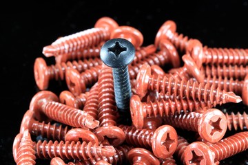 Pile of small red metal screws with one black screw in middle on black background - Individuality...