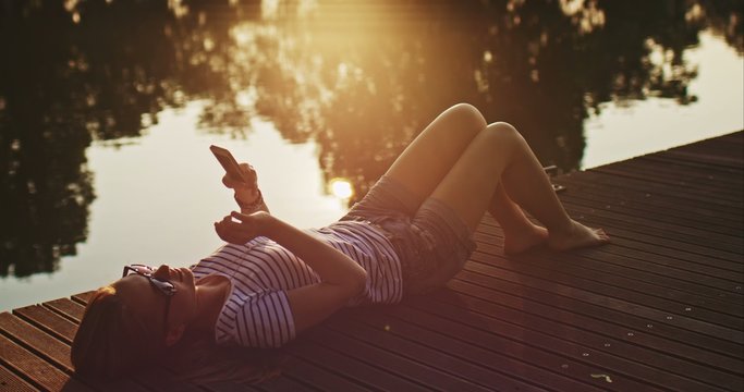 Woman using smartphone outdoors, smiling and relaxing on a wooden jetty near the lake with sunny backlit background, lens flare.  4K, DCi resolution, slow motion. 