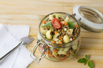 Jar with salad of vegetables, pasta and lentils on a wooden surface