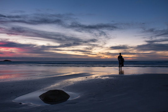 Surfer standing on an isolated beach watching a beautiful sunset