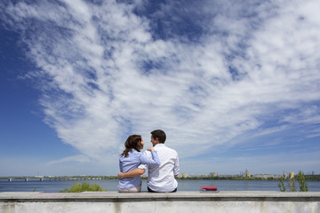 Young couple and cloudy sky.
Two people male and female sitting on railing embracing in front of urban view with river and blue sky clouds
