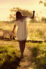 Young girl goes barefoot on a dirt road in a field at sunset