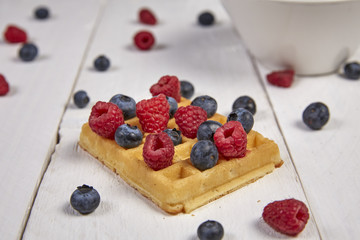 Different berries on a waffle and in a bowl on a white wooden table