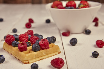 Different berries on a waffle and in a bowl on a white wooden table