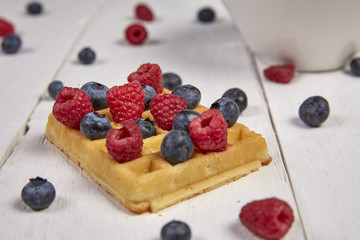 Different berries on a waffle and in a bowl on a white wooden table