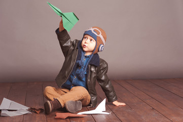 Little boy 4-5 year old playing with paper planes in room. Wearing stylish leather jacket and knitted pilot hat over gray. 