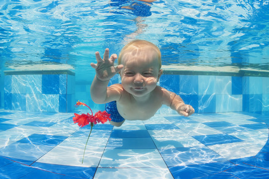 Smiling baby boy diving underwater with fun for red flower in blue pool Active lifestyle, child swimming lesson with parents, and water sports activity during family summer vacation in tropical resort