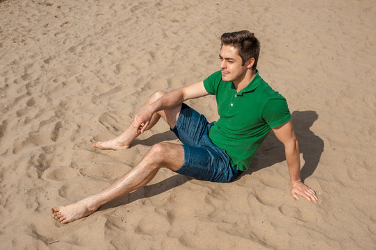 Young fit man wearing green polo sits on a sand at the beach on sunny day