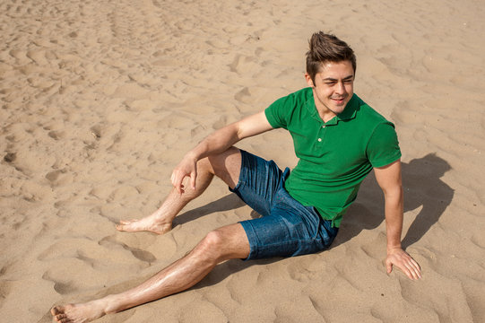Young fit man wearing green polo sits on a sand at the beach on sunny day