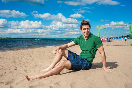 Young fit man wearing green polo sits on a sand at the beach on sunny day under scenic blue cloudy sky