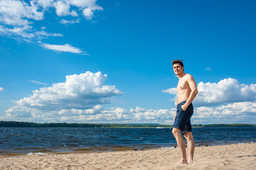 Fit young man with bare torso standing at the beach under scenic blue sky on sunny day