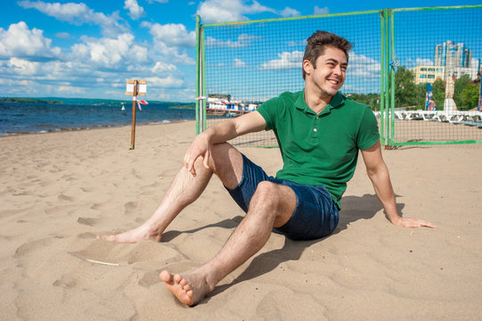 Young fit man wearing green polo sits on a sand at the beach on sunny day under scenic blue cloudy sky