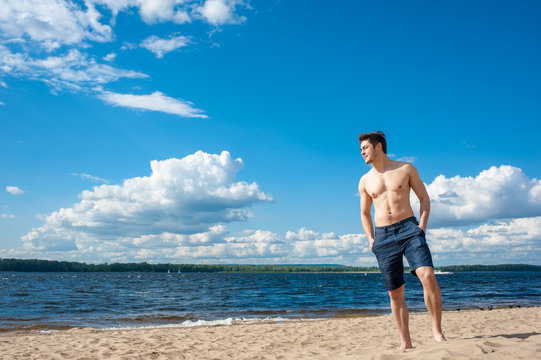 Fit young man with bare torso standing at the beach under scenic blue sky on sunny day