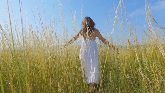 Beautiful young woman with a long white dress walking through long grass and gently touching the stems.