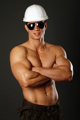 Smiling muscular builder in sunglasses with a protective helmet on gray background