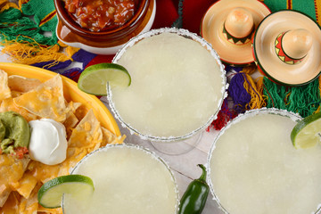 Margaritas: High angle view of three margarita cocktails surrounded by nachos, chips and salsa on a bright Mexican, table cloth. Horizontal format. Perfect for Cinco de Mayo projects. - obrazy, fototapety, plakaty