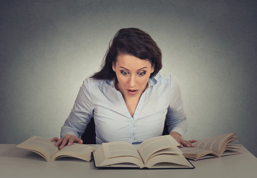 shocked woman sitting at desk with many opened books reading