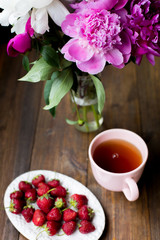 white and pink peonies, black tea and fresh strawberries from farmers market on a dark wood table in rustic style