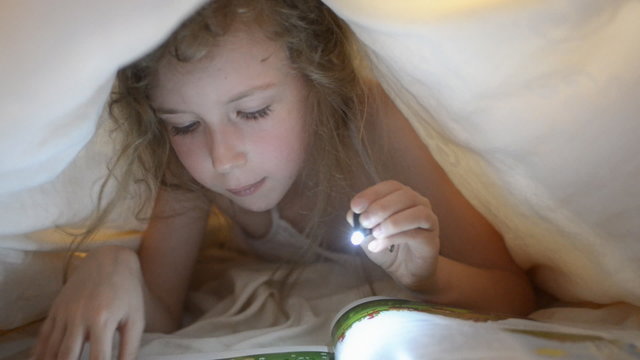 Little girl reading book under the covers.