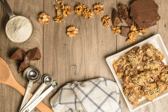 Overhead view of homemade chocolate walnut cookies with ingredients and kitchen utensils on a light rustic wood table. Open space in center for logo or text.