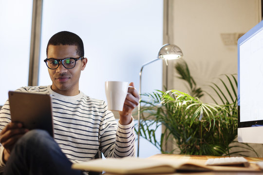 Young creative man with mini tablet having coffee break in his home office
