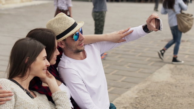 Young friends taking selfie photo with cellphone sitting on bench in city
