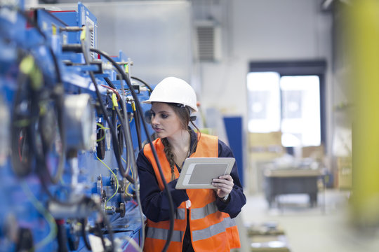 Technician with reflective vest in factory hall inspecting machine with digital tablet