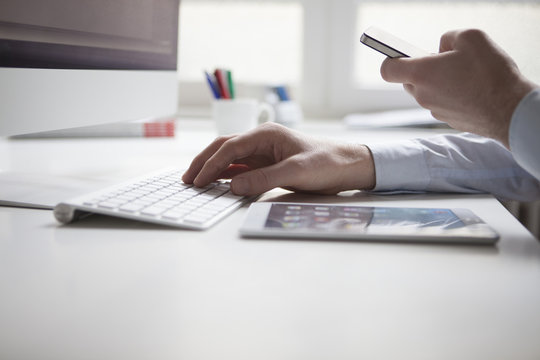Man's hand typing on keyboard of computer while using smartphone