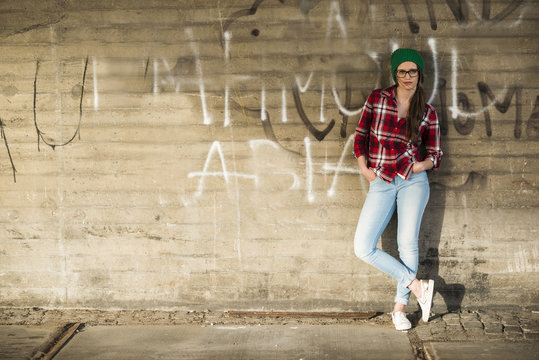 Young woman with checkered shirt and wooly hat leaning at graffiti wall