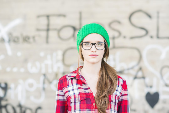 Young woman with checkered shirt and green wooly hat at graffiti wall