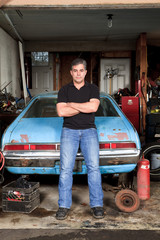 A man leaning against his old car in his messy garage.