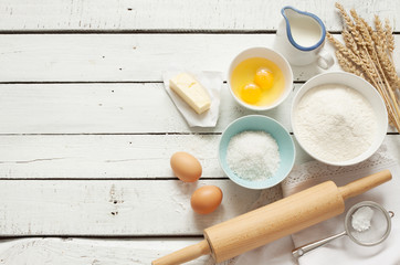 Baking cake in rustic kitchen - dough recipe ingredients (eggs, flour, milk, butter, sugar) on white planked wooden table from above. Background layout with free text space.