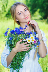 Young charming woman holding flower bouquet with camomiles and cornflowers  and smiling, against green summer nature