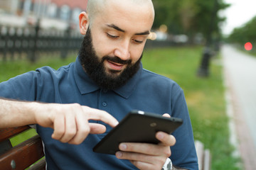 Fototapeta premium Bearded bald man sitting on a bench and read tablet at the alley