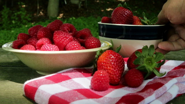 raspberries and strawberry on table and hand is taking out bowl with strawberries and returning back to table
