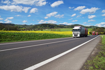 Trucks driving on the asphalt road around the yellow flowering rapeseed field in rural landscape