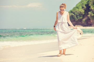 Fototapeta na wymiar Young woman in long white dress holding hat walks along tropical beach having great summer time on holidays