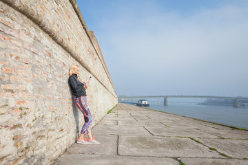 Woman resting after morning workout leaning on the wall and using mobile phone