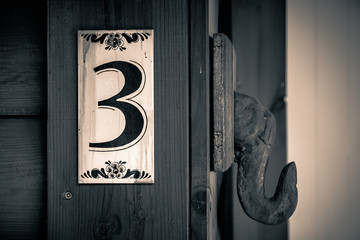 Old apartment number sign on the wall of wood with the number three on it.
Sepia colors.
