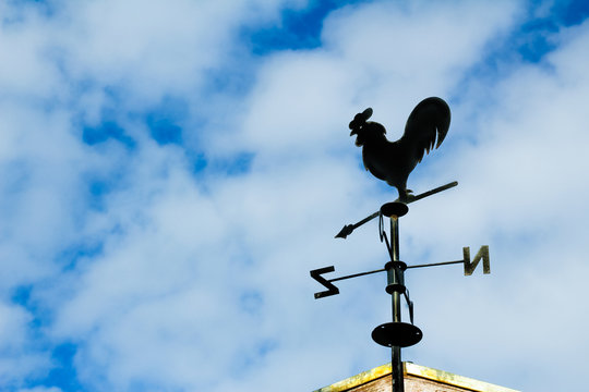Black weathervane in the form of a rooster.
The background has a blue sky with clouds.