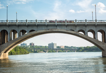 horizontal image of an arched concrete bridge framing skyscrapers and trees in the background with a river flowing underneath and a blue sky in the summer time