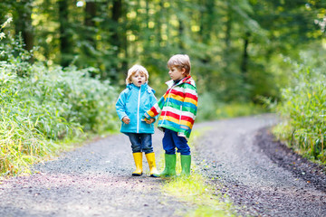 Obraz premium Two little sibling boys in colorful raincoats and boots walking
